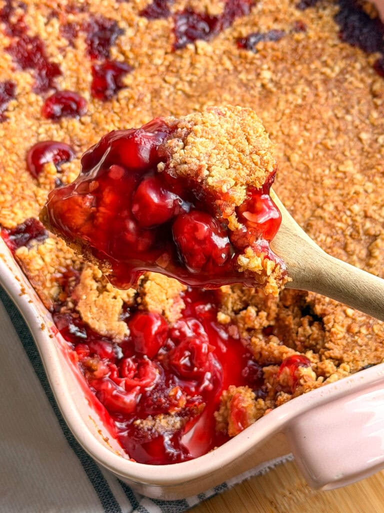 A wooden spoon lifts a scoop of cherry crisp from a pink baking dish filled with red fruit filling and a golden brown crumb topping.