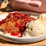 A serving of cherry crumble topped with two scoops of vanilla ice cream on a white plate, with a pink baking dish in the background.