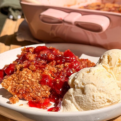 A serving of cherry crumble topped with two scoops of vanilla ice cream on a white plate, with a pink baking dish in the background.