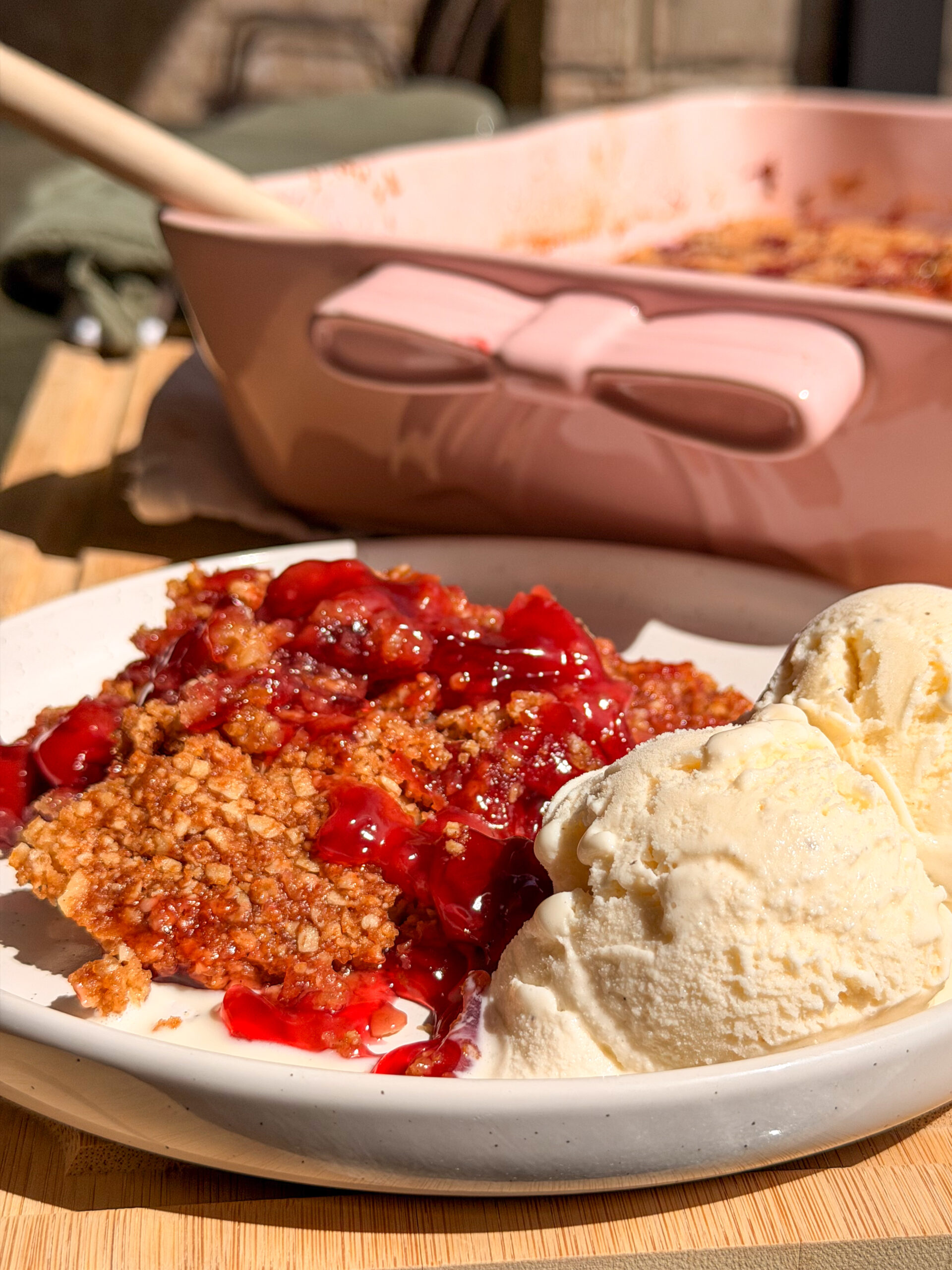 A serving of cherry crumble topped with two scoops of vanilla ice cream on a white plate, with a pink baking dish in the background.