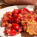 A serving of cherry crisp topped with a scoop of ice cream on a white plate, with a pink baking dish in the background.