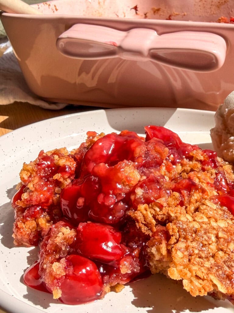 A serving of cherry crisp topped with a scoop of ice cream on a white plate, with a pink baking dish in the background.