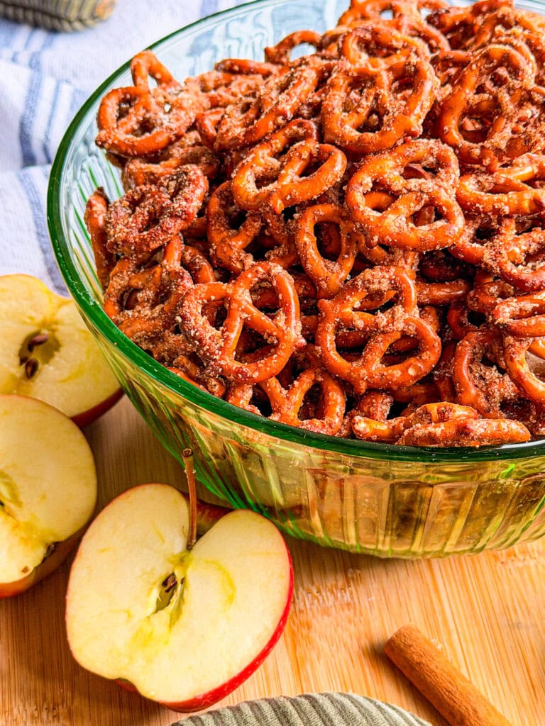 A green glass bowl filled with cinnamon-sugar pretzels, surrounded by apple slices and a cinnamon stick on a wooden surface.