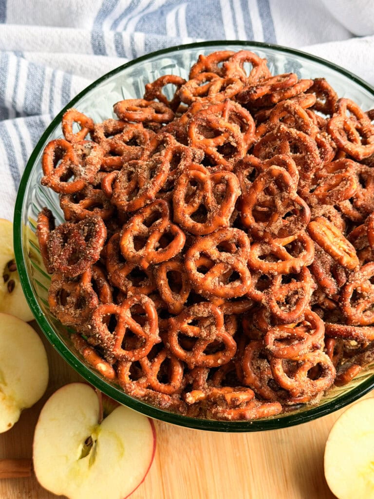 A glass bowl filled with cinnamon-sugar coated pretzels, sitting on a wooden cutting board with sliced apples.