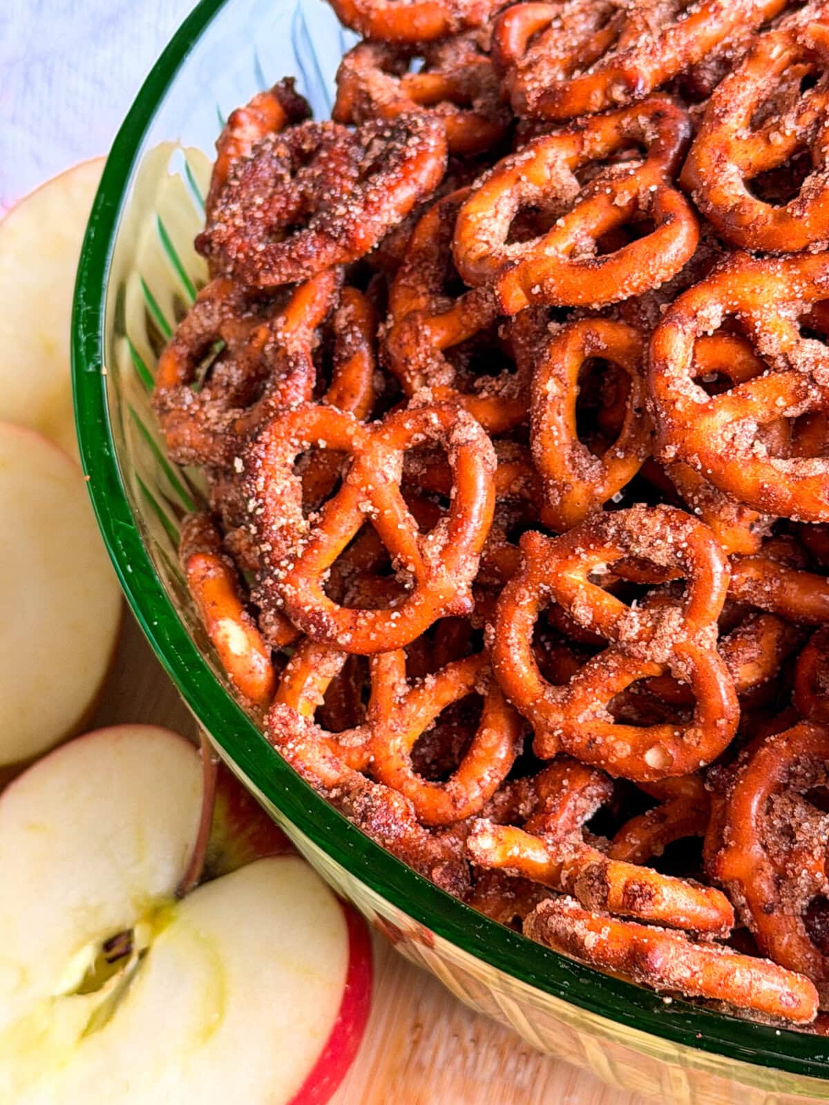A glass bowl filled with cinnamon-sugar coated pretzels, sitting next to sliced apples.