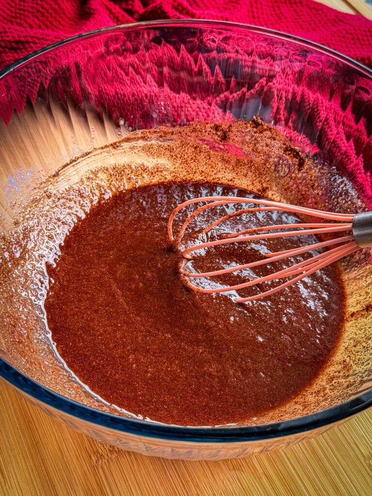 A whisk sits in a glass bowl filled with smooth, dark brown liquid batter.