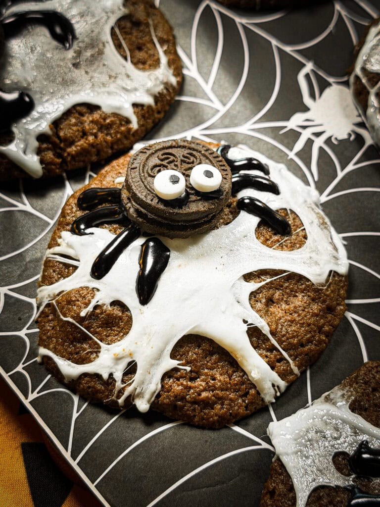 A chocolate cookie decorated with white icing spiderwebs and a sandwich cookie spider with candy eyes.