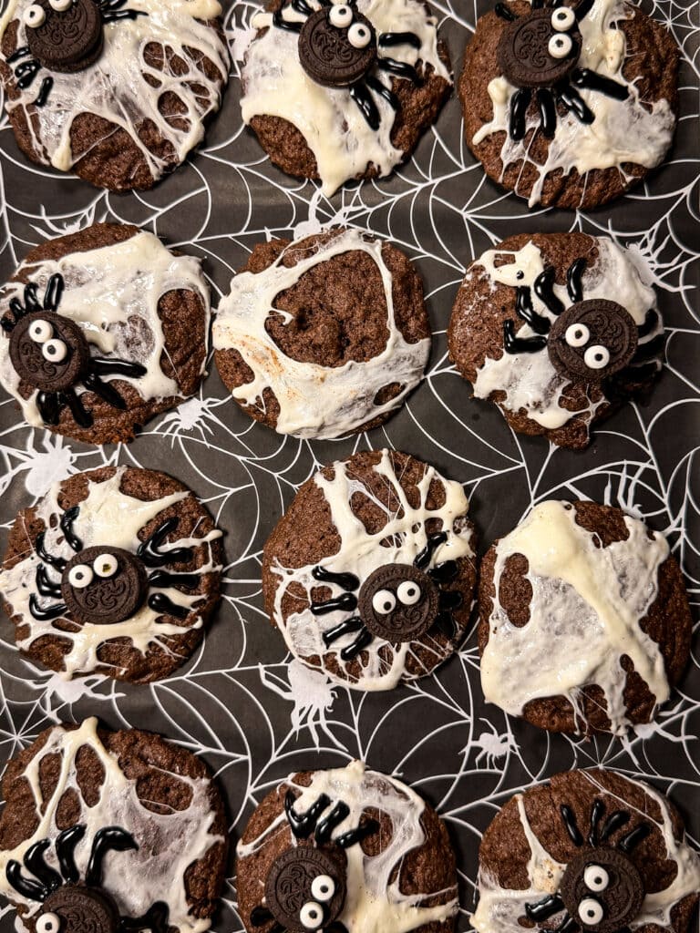 Nine chocolate cookies arranged on black paper with a spiderweb print, decorated with white icing webs and Oreo spider toppers.