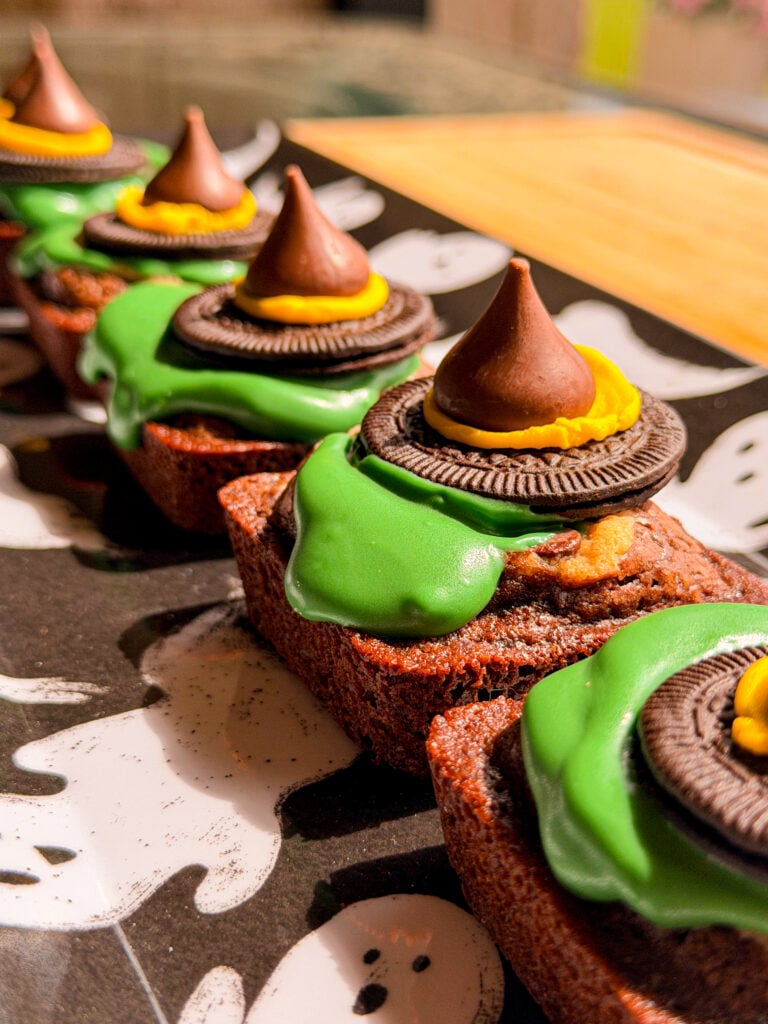 Small rectangular brownies decorated as witch hats, featuring green icing, a sandwich cookie, and a chocolate kiss on top, arranged on a ghost-patterned tray.