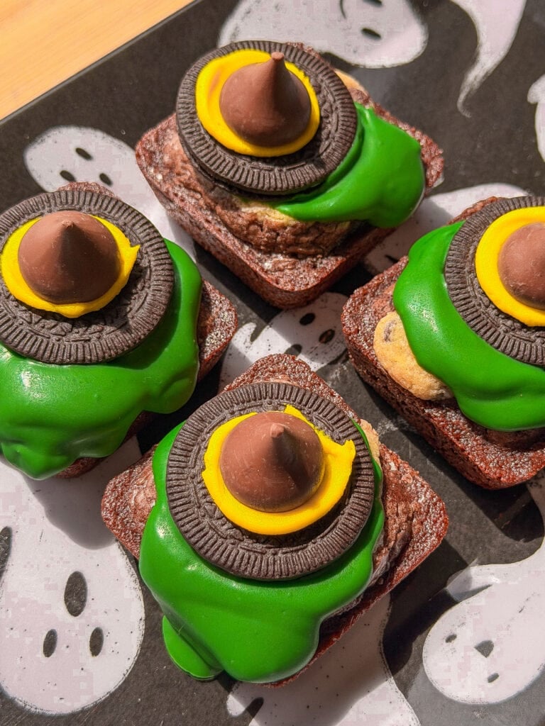 Four brownie bites decorated as witch hats with green icing, cookie bases, and chocolate kiss tops, arranged on a plate with ghost patterns.