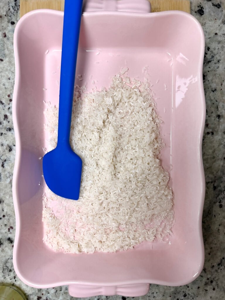 A pile of raw white rice in a rectangular pink baking dish with a blue silicone spatula.