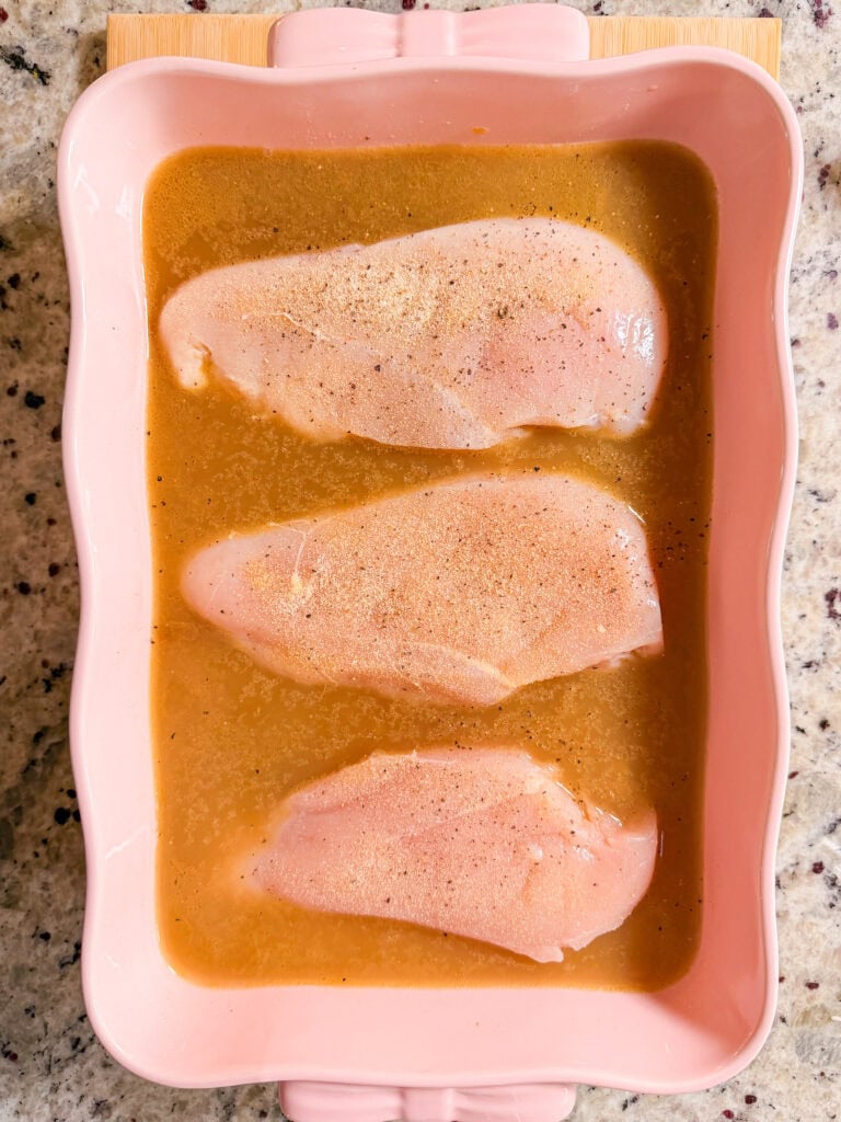Three raw chicken breasts seasoned with spices and resting in a light brown marinade inside a pink ceramic baking dish.