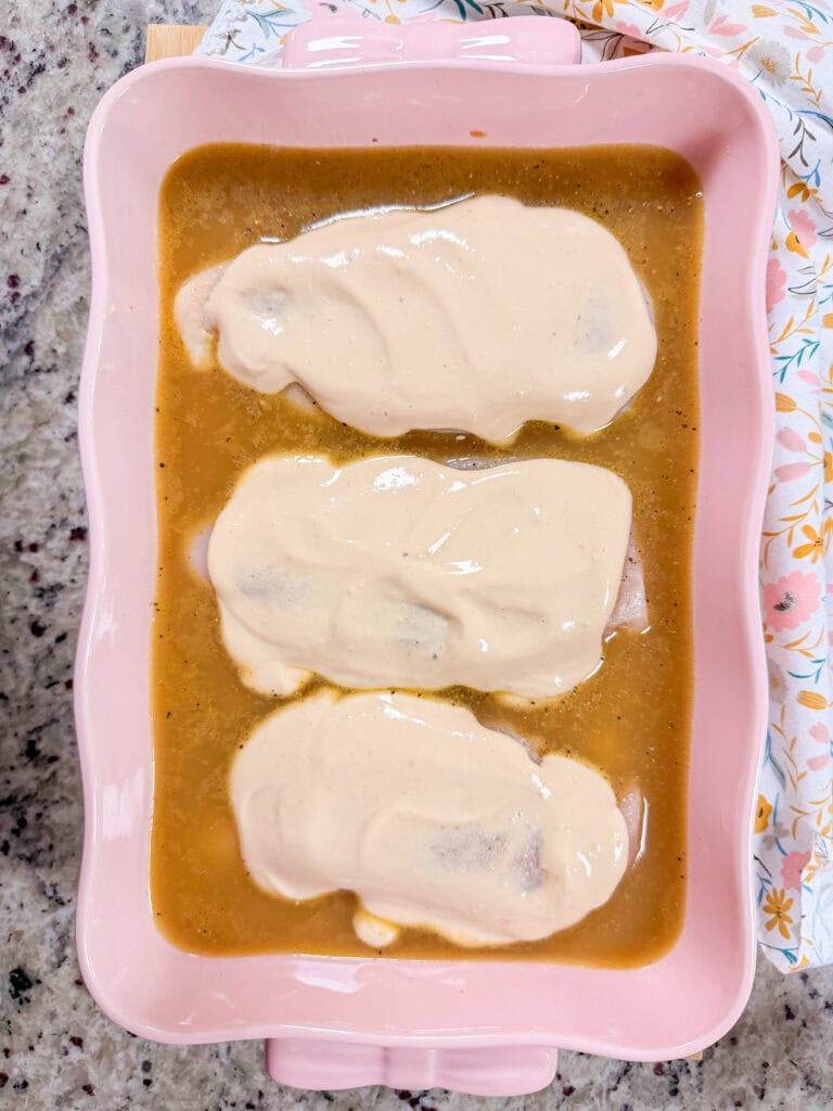 Three chicken breasts covered in a creamy sauce and brown gravy inside a pink rectangular baking dish.