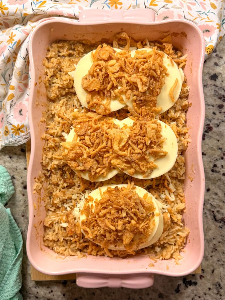 A rectangular pink baking dish containing rice casserole topped with three slices of cheese and fried onions.