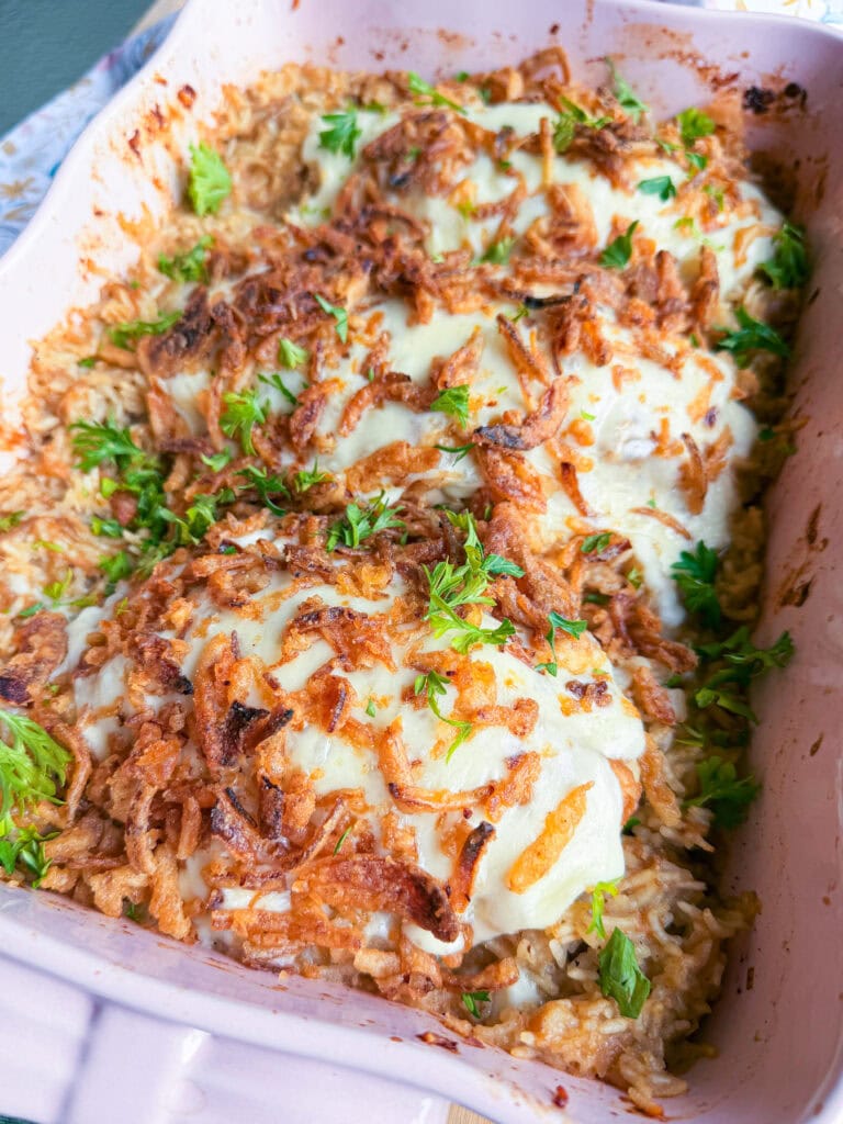 Casserole dish containing chicken topped with melted cheese, crispy fried onions, and fresh parsley, served over a bed of rice.