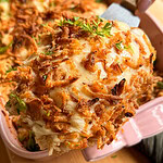 A spatula lifts a serving of cheesy casserole topped with crispy fried onions and parsley from a pink baking dish.
