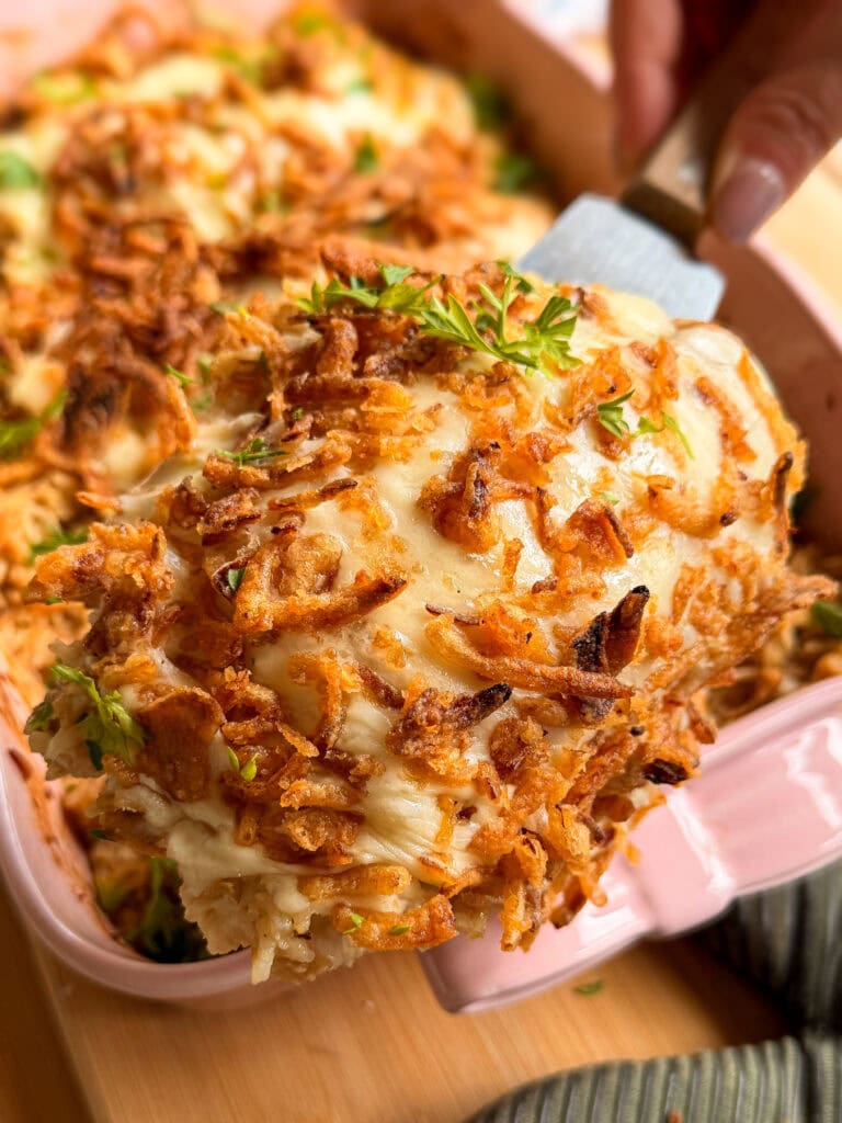 A spatula lifts a serving of cheesy casserole topped with crispy fried onions and parsley from a pink baking dish.