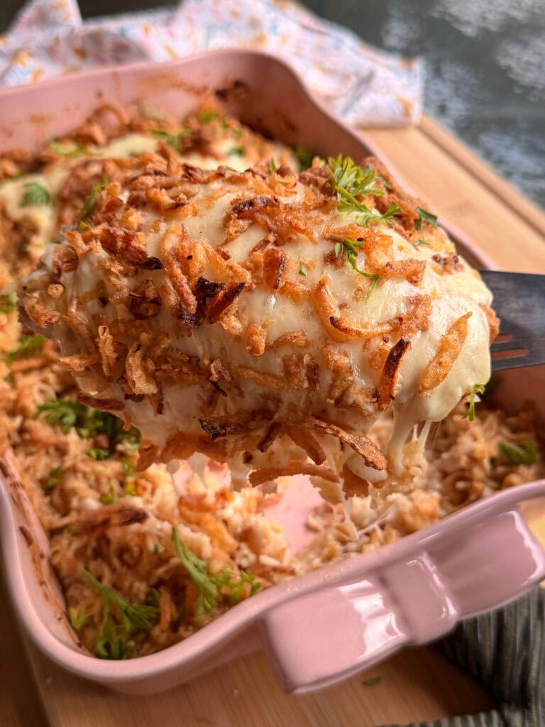 A serving of creamy chicken casserole topped with melted cheese and crispy fried onions being lifted from a pink baking dish with a spatula.