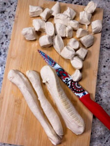 A patterned knife sits on a wooden cutting board next to several ropes of raw dough and cut dough pieces.
