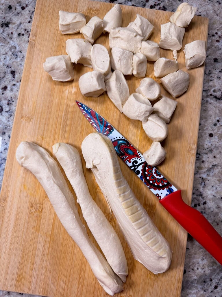 A patterned knife sits on a wooden cutting board next to several ropes of raw dough and cut dough pieces.