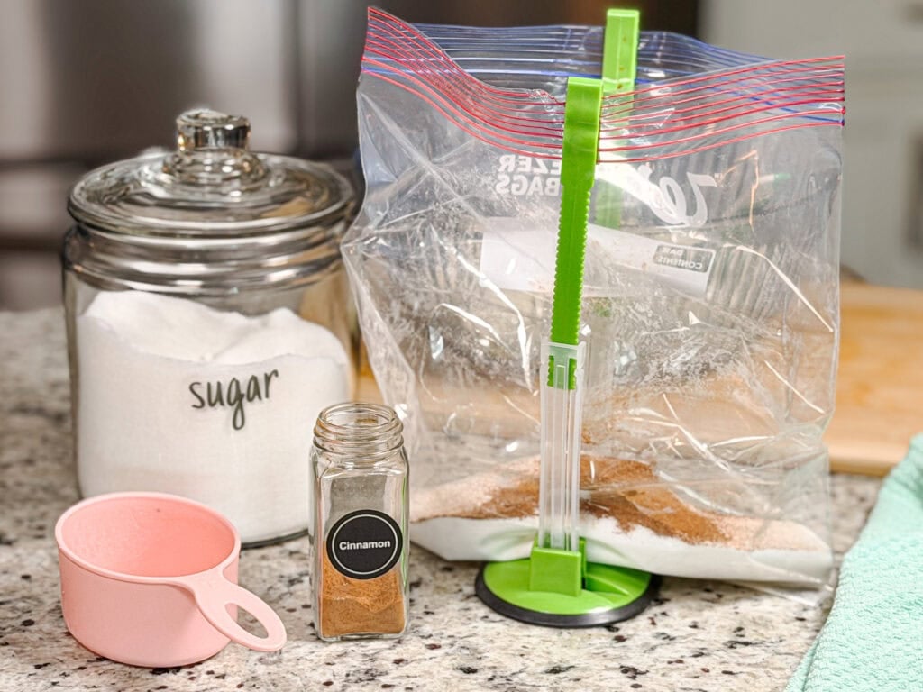 A jar of sugar, a pink measuring cup, a jar of cinnamon, and a plastic freezer bag held open by a green stand on a kitchen counter.