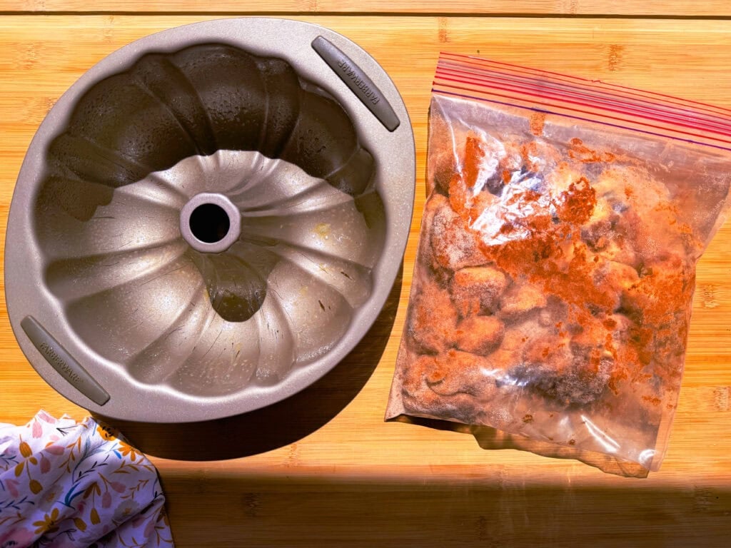 A metal fluted bundt pan sits next to a plastic storage bag filled with frozen, orange-dusted food on a wooden surface.