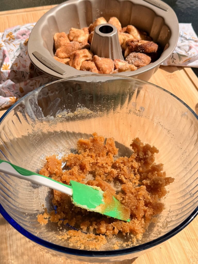 A glass bowl containing a brown sugar and cinnamon mixture with a green spatula, and a Bundt pan filled with coated dough pieces in the background.