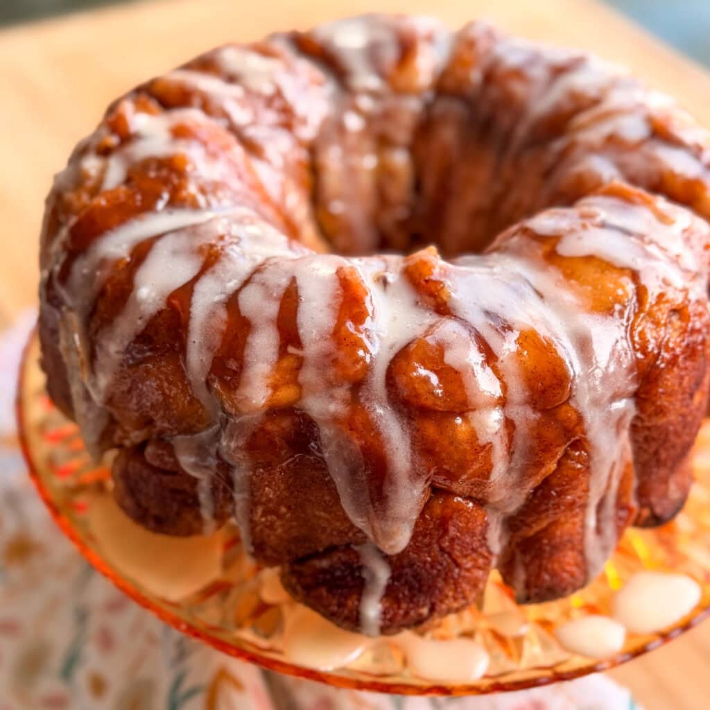 A circular monkey bread ring drizzled with white icing on a glass pedestal cake stand.