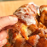 A hand pulls a glazed, cinnamon-dusted piece from a monkey bread loaf on a glass plate.