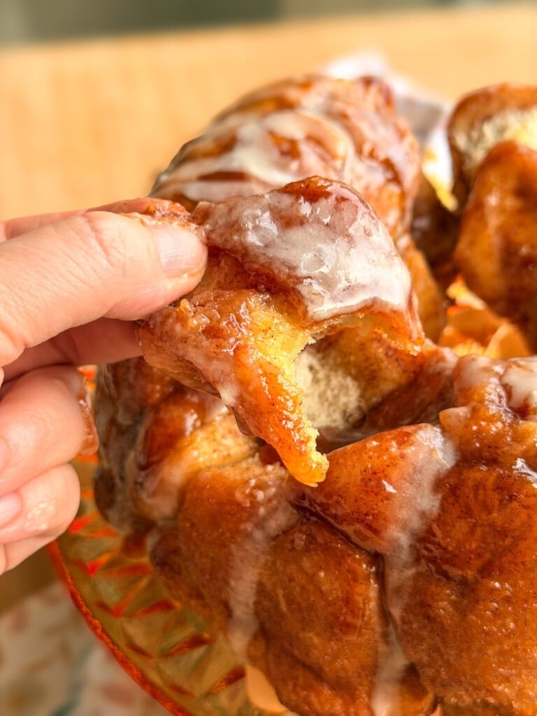A hand pulls a glazed, cinnamon-dusted piece from a monkey bread loaf on a glass plate.
