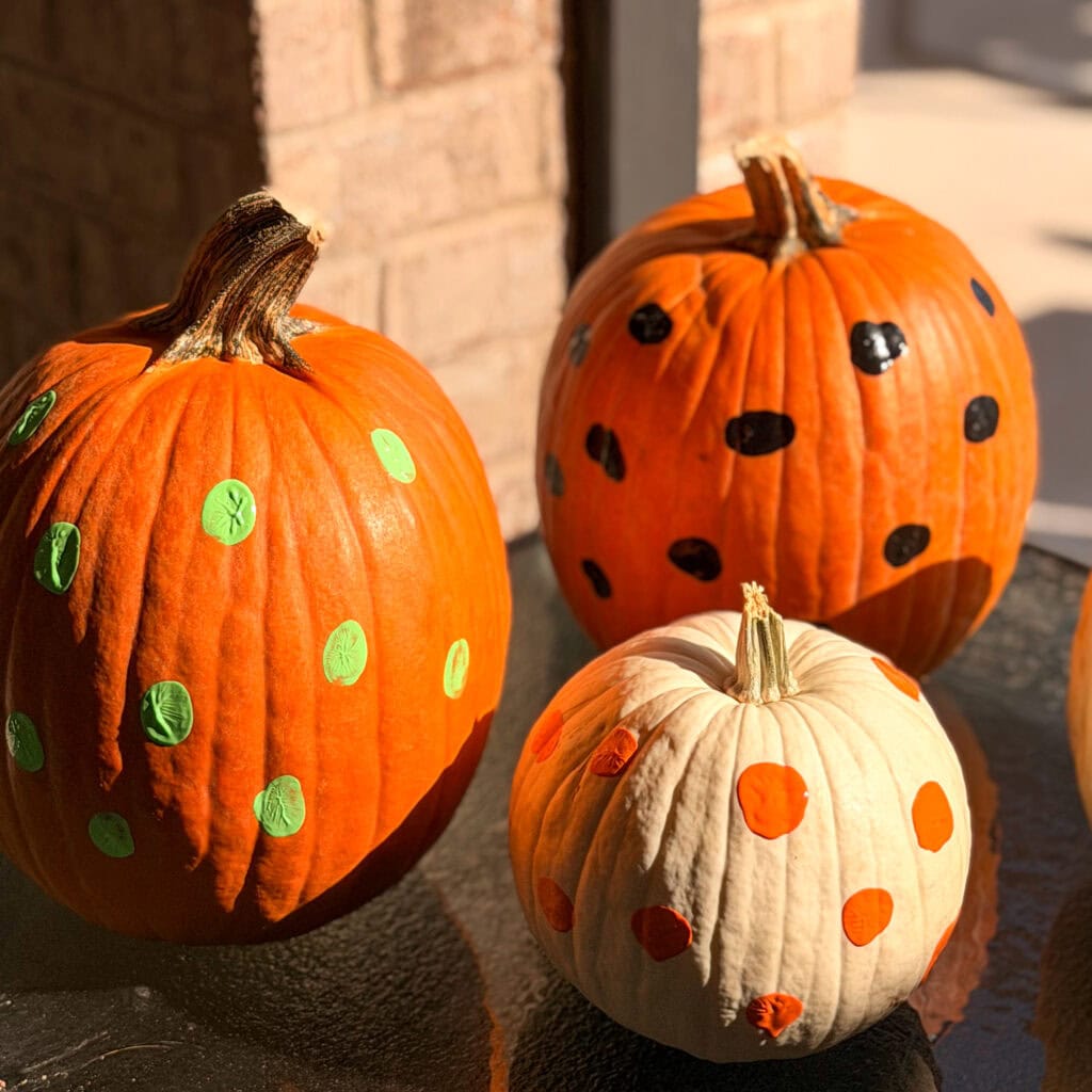 Two orange pumpkins with green and black polka dots and one white pumpkin with orange polka dots sitting on a glass table.