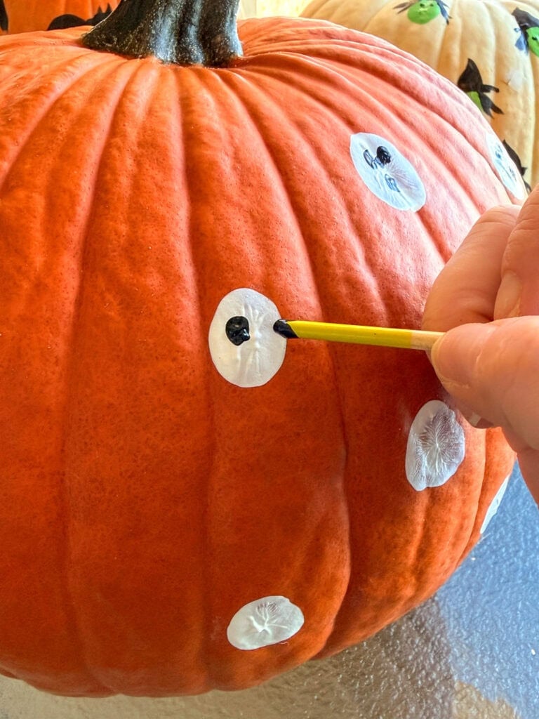 A hand uses a yellow paintbrush to add a black dot to a white circle painted on a pumpkin.