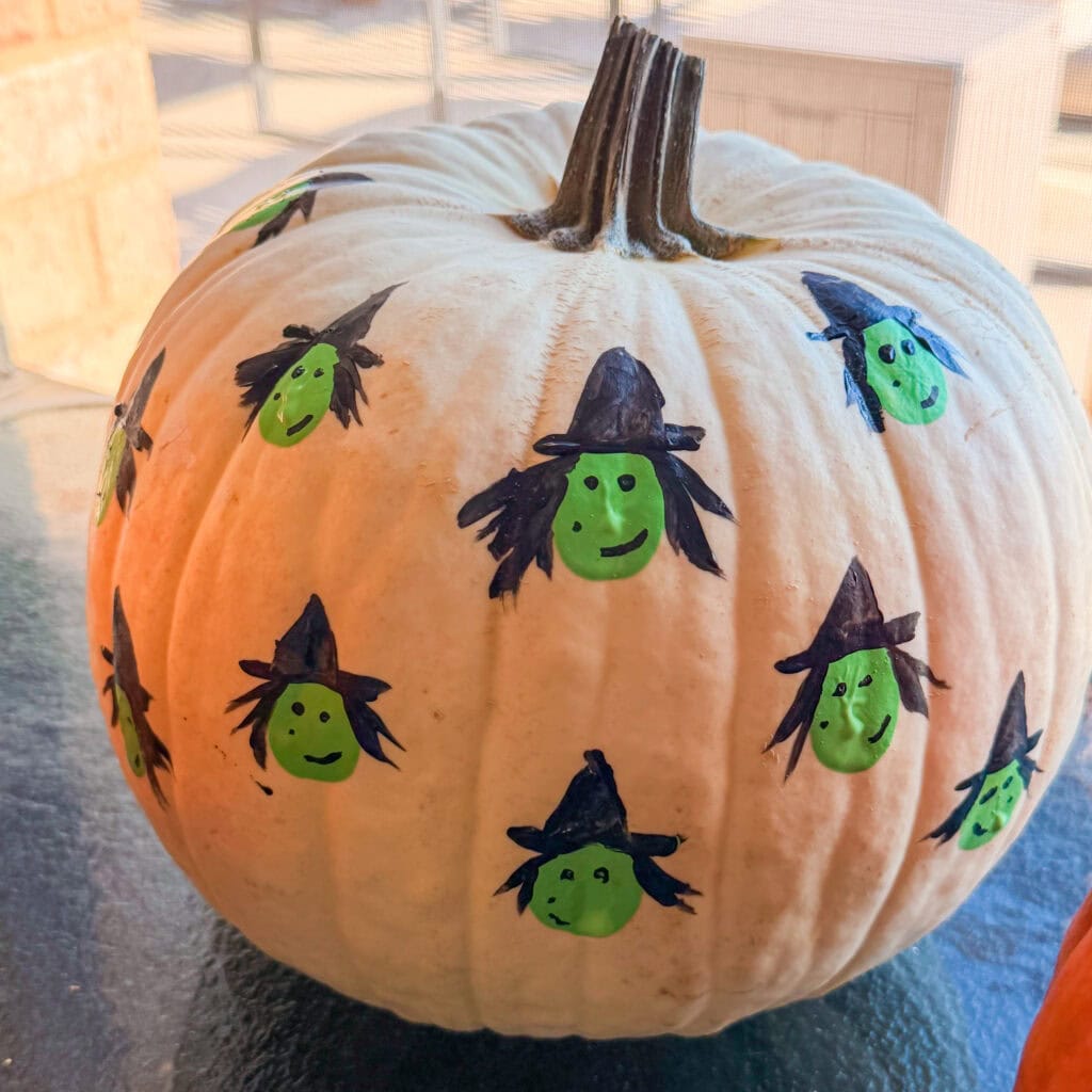 A white pumpkin decorated with several hand-drawn images of green-faced witches wearing black hats.