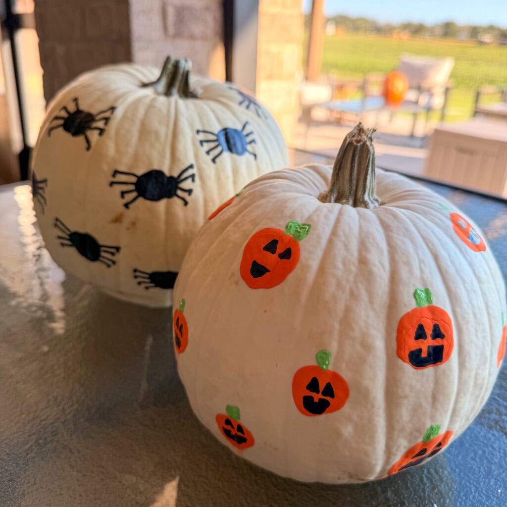 Two white pumpkins on a table, one decorated with painted black spiders and the other with painted orange jack-o-lanterns.
