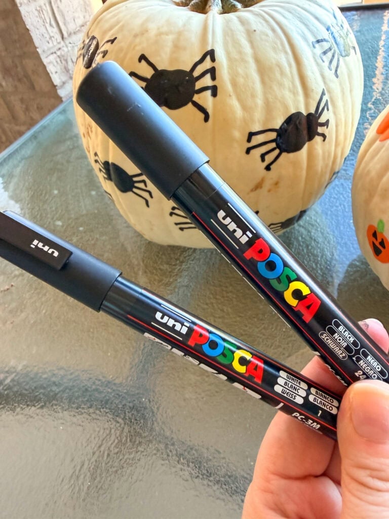 A hand holding two black Uni Posca paint markers, one labeled white and one labeled black, in front of a white pumpkin decorated with spider drawings.