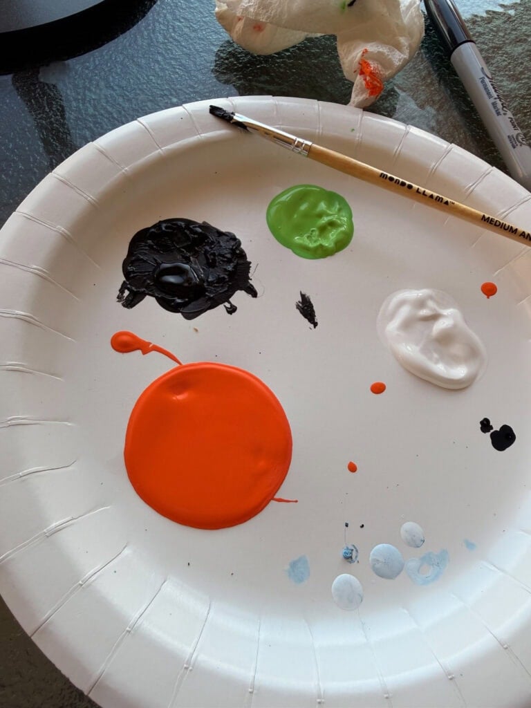A white paper plate used as a paint palette, featuring dabs of orange, black, green, and white paint, with a paintbrush resting on the edge.