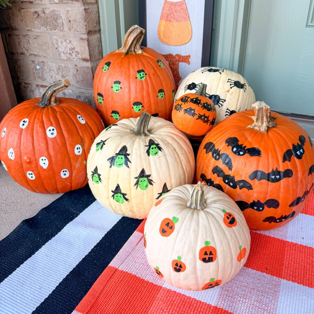 A group of orange and white pumpkins painted with various Halloween designs, including ghosts, witches, bats, spiders, and jack-o-lanterns, arranged on a striped rug in front of a door.