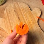 A hand holds a carrot slice with a V-shaped notch cut into the top, positioned over a wooden cutting board.