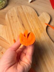 A hand holds a carrot slice with a V-shaped notch cut into the top, positioned over a wooden cutting board.