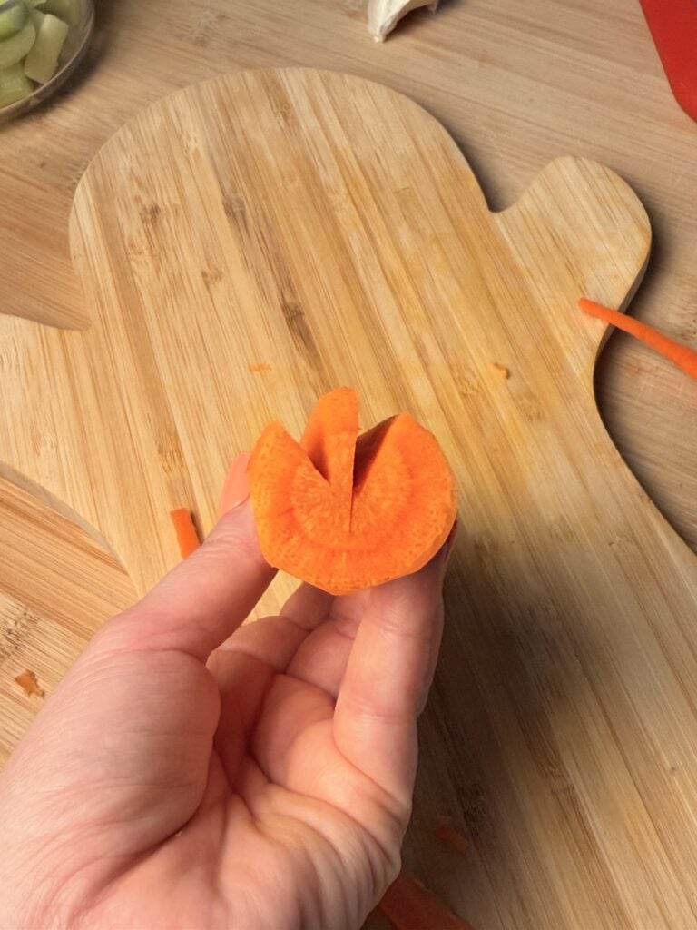 A hand holds a carrot slice with a V-shaped notch cut into the top, positioned over a wooden cutting board.
