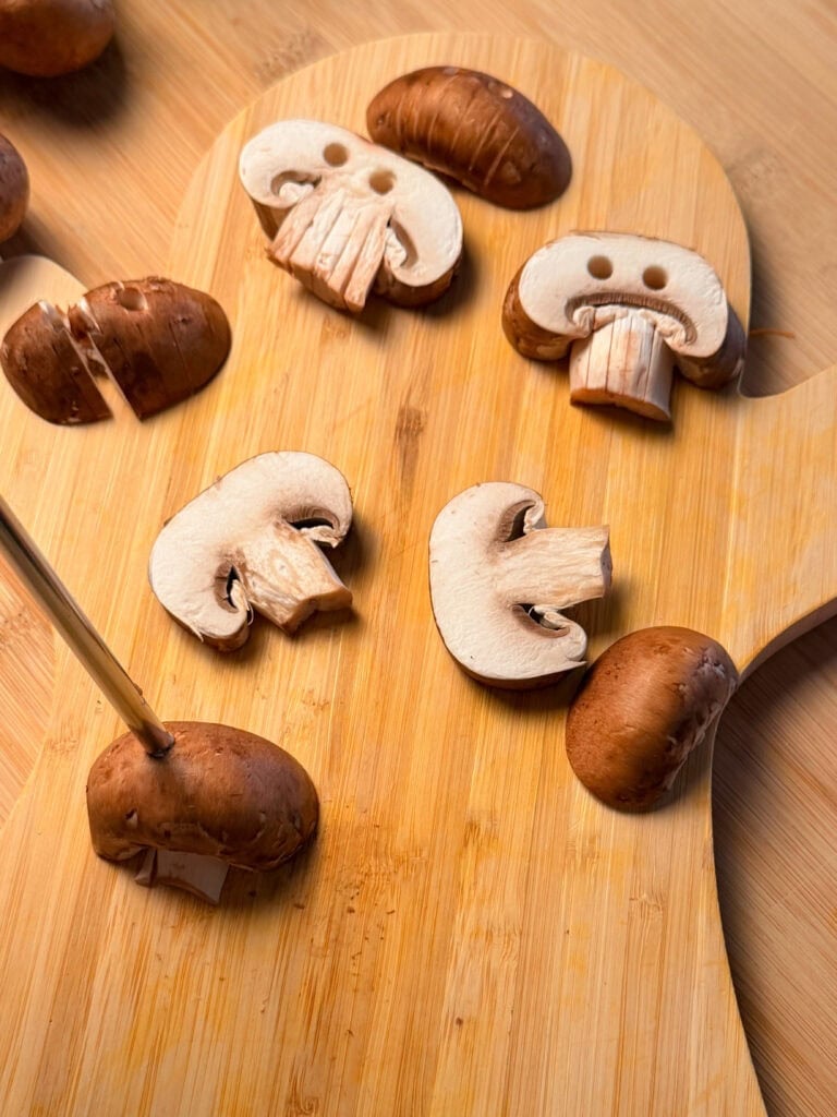 Cremini mushroom slices arranged on a wooden cutting board, with two slices positioned to resemble faces.