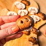 A hand holds a brown mushroom carved to look like a skull, with several other skull-carved mushrooms on a wooden cutting board nearby.
