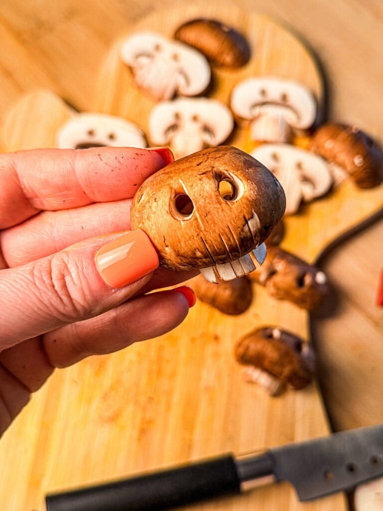 A hand holds a brown mushroom carved to look like a skull, with several other skull-carved mushrooms on a wooden cutting board nearby.