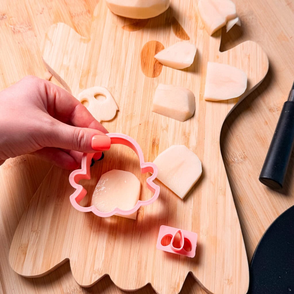 A hand presses a pink ghost-shaped cookie cutter into a slice of potato on a ghost-shaped wooden cutting board.
