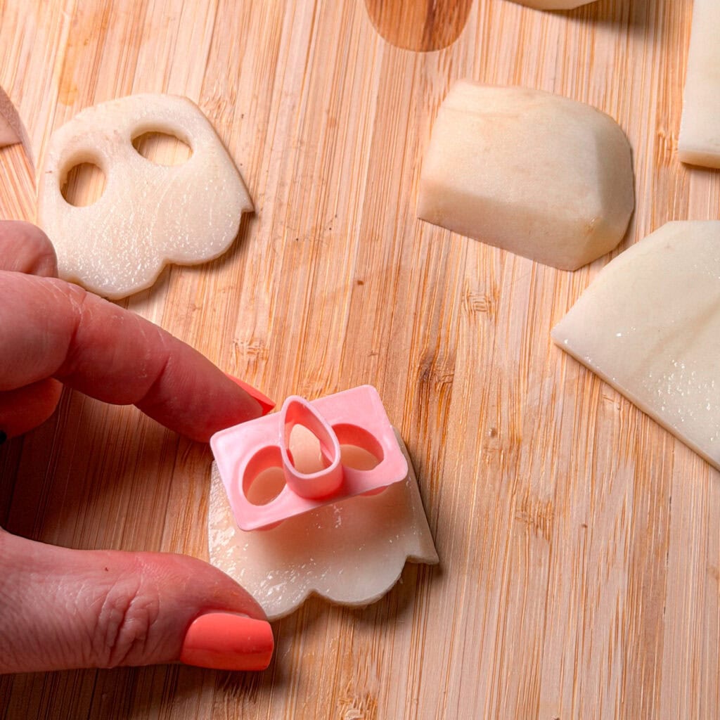 A hand uses a pink plastic ghost-shaped cutter on a slice of raw potato on a wooden cutting board.