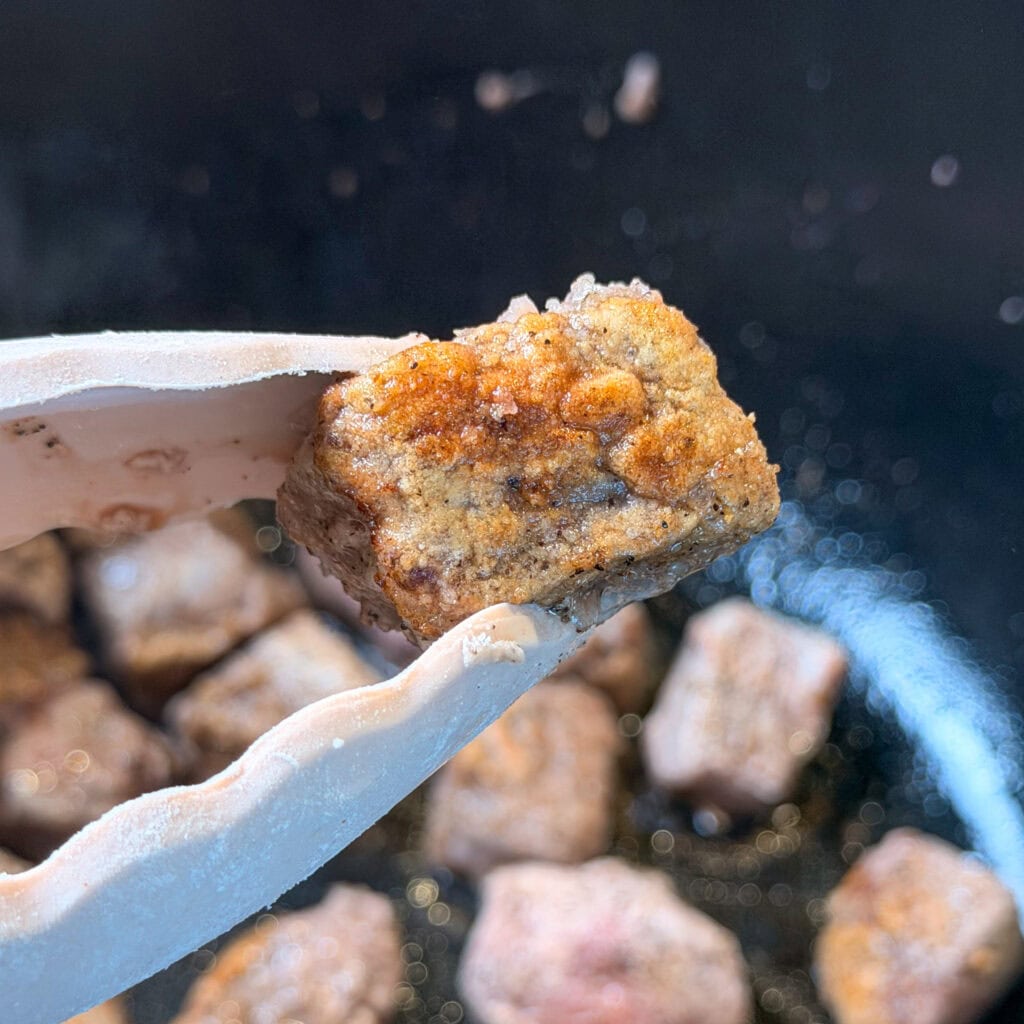 Tongs holding a seared, bite-sized cube of seasoned meat over a pan of cooking meat.