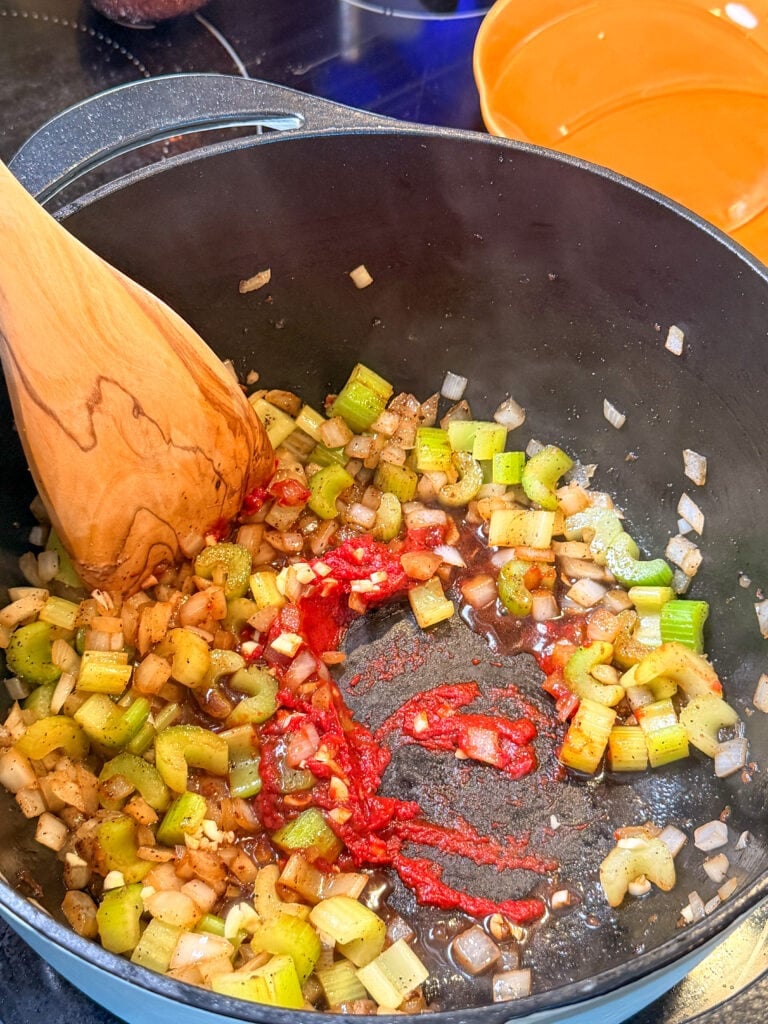 A wooden spoon stirs sautéed diced celery and onions with a dollop of red tomato paste in a black pot.