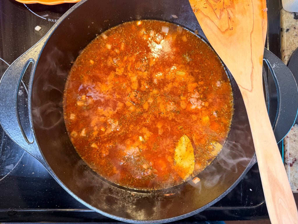 A steaming red stew with a bay leaf simmering in a black cast iron pot, with a wooden spatula resting on the edge.