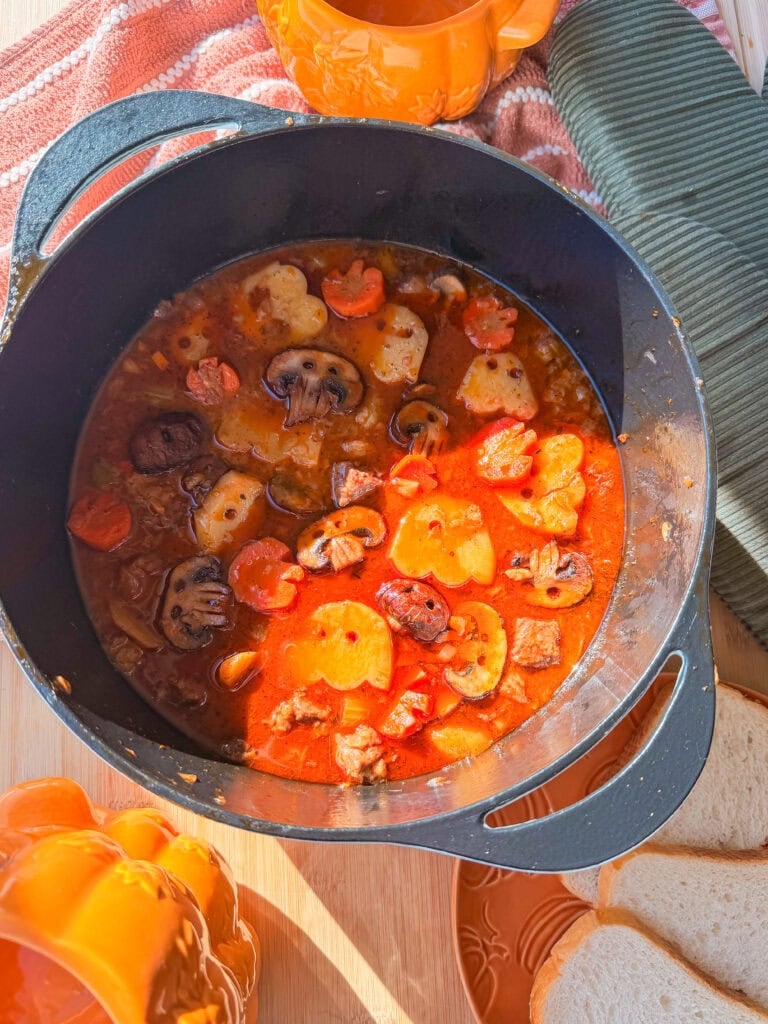 A black Dutch oven filled with a hearty stew containing sliced mushrooms, carrots, and potatoes, positioned on a table with pumpkin-shaped dishes and sliced bread.