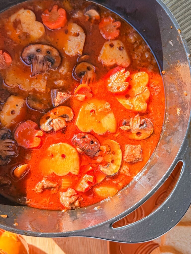 A Dutch oven filled with a rich, red stew containing sliced carrots, potatoes, mushrooms, and pieces of meat.